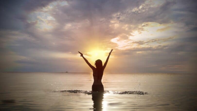 Imagem da silhueta de uma mulher de cabelos curtos, dentro da água do mar. Ela está de braços abertos, feliz, olhando para cima e recebendo a energia que vem do sol, simbolizando o conceito de luz interior.