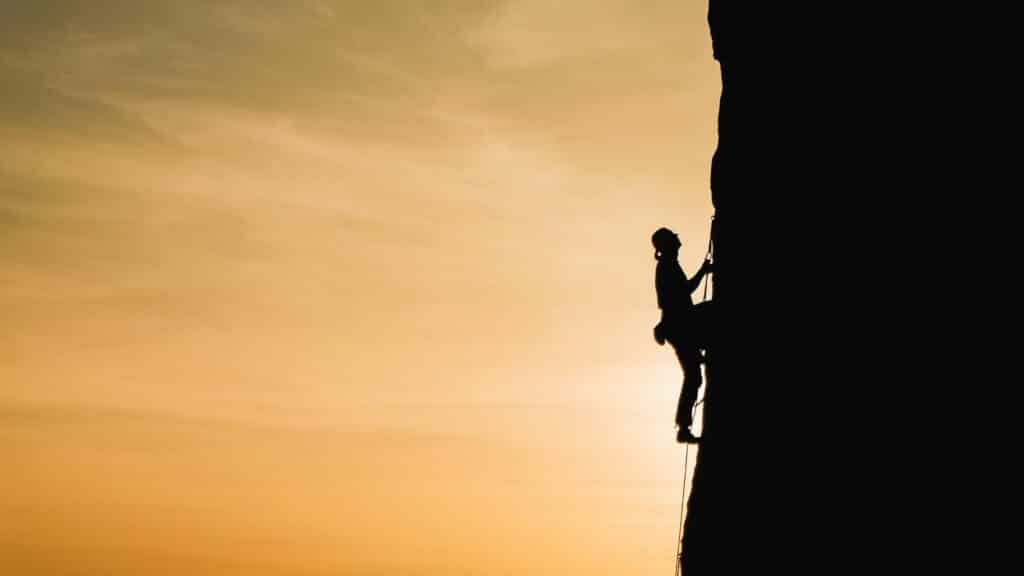 Imagem da silhueta de um homem escalando uma montanha em um lindo final de tarde, simbolizando os desafios e obstáculos que temos que enfrentar.
