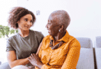 Uma avó e uma neta adulta estão sentadas em um sofá. Elas conversam e sorriem uma para a outra.