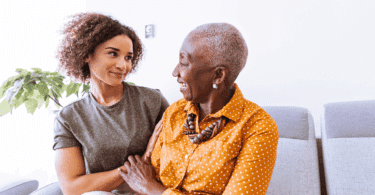 Uma avó e uma neta adulta estão sentadas em um sofá. Elas conversam e sorriem uma para a outra.