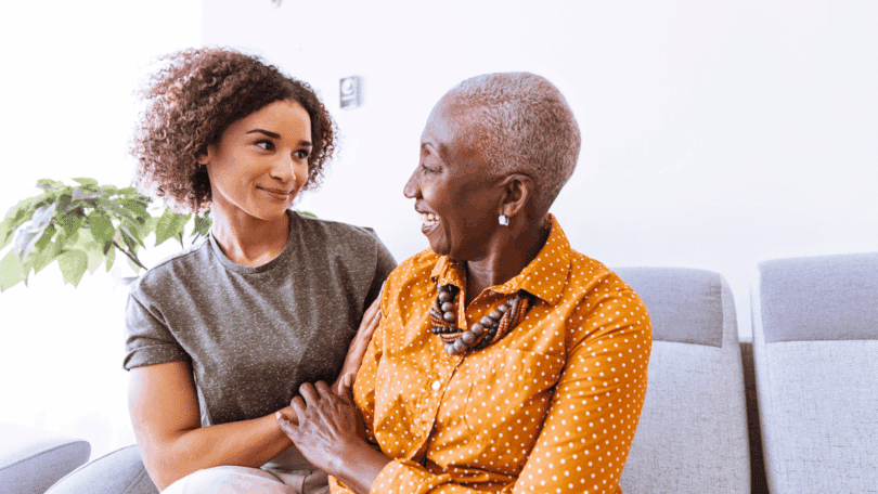 Uma avó e uma neta adulta estão sentadas em um sofá. Elas conversam e sorriem uma para a outra.