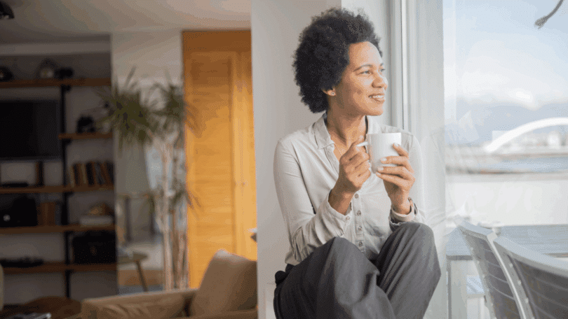 Uma mulher está sentada perto da janela da sala da sua casa. Ela olha pela janela, contempla a paisagem lá fora e toma uma xícara de café.