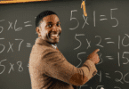 Um professor de matemática está ensinando o conteúdo que está na lousa. Ele sorri e aponta para a lousa.
