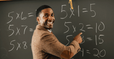 Um professor de matemática está ensinando o conteúdo que está na lousa. Ele sorri e aponta para a lousa.