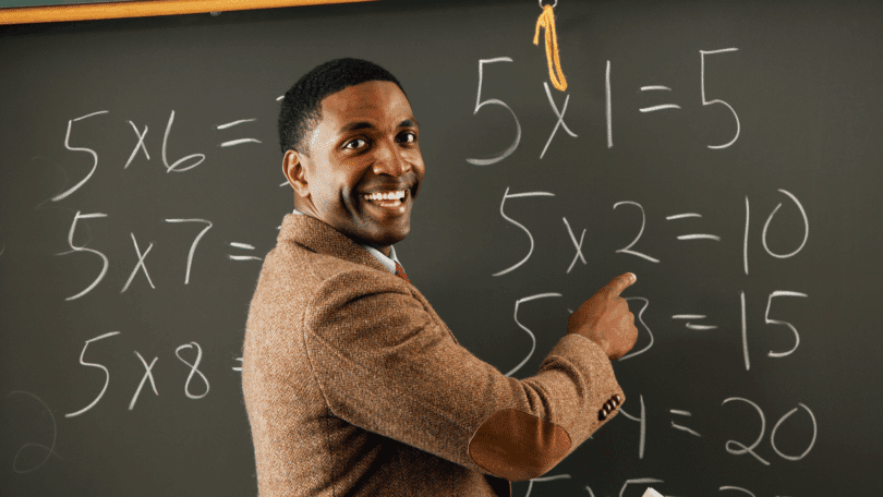 Um professor de matemática está ensinando o conteúdo que está na lousa. Ele sorri e aponta para a lousa.