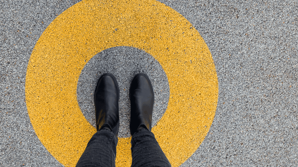 Os pés de uma pessoa que usa botas pretas estão em destaque na imagem. A pessoa está em pé em uma rua asfaltada e, em volta dela, há um círculo amarelo, indicando que ela está presa na sua zona de conforto.