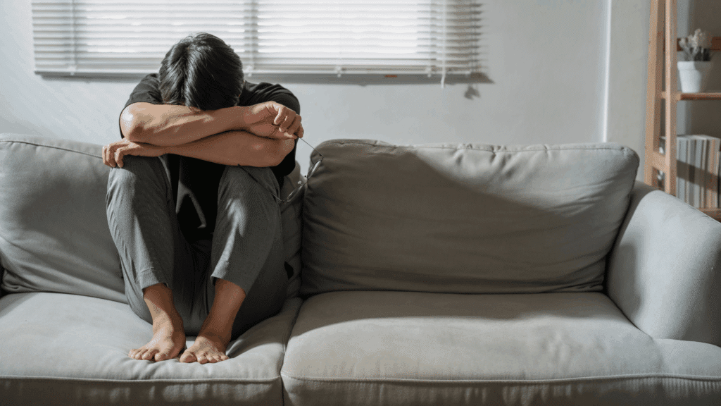 Um homem adulto está triste. Ele está sentado no sofá da sua casa cabisbaixo, com a cabeça inclinada para baixo e apoiando-a nos braços.