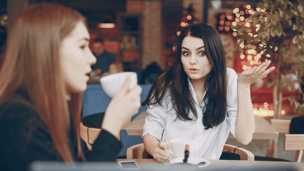 Duas mulheres sentadas em um café, conversando enquanto tomam café; uma delas parece surpresa ou confusa, com expressão de espanto e a mão levantada, enquanto a outra fala segurando uma xícara.