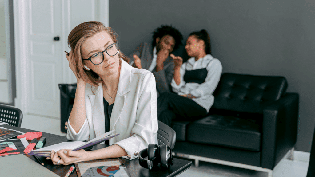 Mulher sentada à mesa de trabalho, com expressão pensativa e olhar desconfortável, enquanto duas pessoas conversam ao fundo em um sofá, aparentemente cochichando.