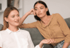 Duas mulheres sentadas em um sofá conversando; uma delas fala com expressão tranquila, enquanto a outra observa com olhar de desconfiança ou impaciência, apoiando a cabeça na mão.