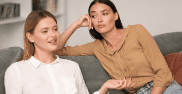 Duas mulheres sentadas em um sofá conversando; uma delas fala com expressão tranquila, enquanto a outra observa com olhar de desconfiança ou impaciência, apoiando a cabeça na mão.