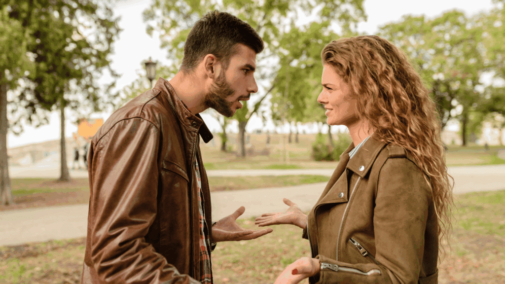Homem e mulher conversando em um parque, de frente um para o outro; o homem parece estar discutindo ou falando com intensidade, enquanto a mulher o observa com expressão calma e ligeiro sorriso.