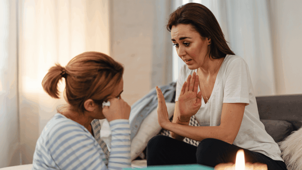 Duas mulheres sentadas em uma sala; uma delas chora segurando um lenço, enquanto a outra, com expressão preocupada, levanta as mãos tentando acalmá-la ou explicar algo.