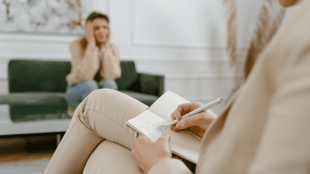 Psicóloga escrevendo em um caderno durante uma sessão de terapia, enquanto outra pessoa ao fundo, sentada em um sofá, aparenta estar angustiada, com as mãos na cabeça.