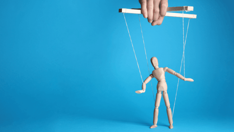Mão segurando fios que controlam um boneco de madeira como uma marionete, sobre um fundo azul.