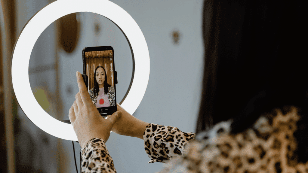 Mulher gravando vídeo com o celular diante de um ring light, usando blusa de estampa animal print, em ambiente interno bem iluminado.