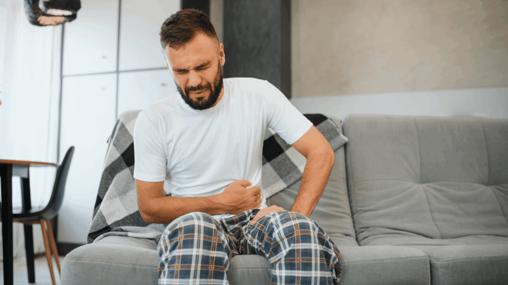 Homem sentado no sofá com expressão de dor, segurando o abdômen, indicando desconforto ou dor abdominal.