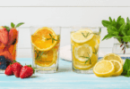 Copos com águas saborizadas de frutas — morango, laranja e limão — decorados com ervas frescas sobre mesa azul, com folhas de hortelã e frutas ao redor.