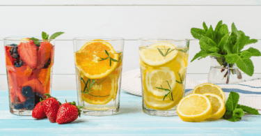 Copos com águas saborizadas de frutas — morango, laranja e limão — decorados com ervas frescas sobre mesa azul, com folhas de hortelã e frutas ao redor.