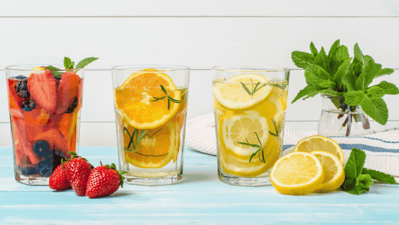 Copos com águas saborizadas de frutas — morango, laranja e limão — decorados com ervas frescas sobre mesa azul, com folhas de hortelã e frutas ao redor.