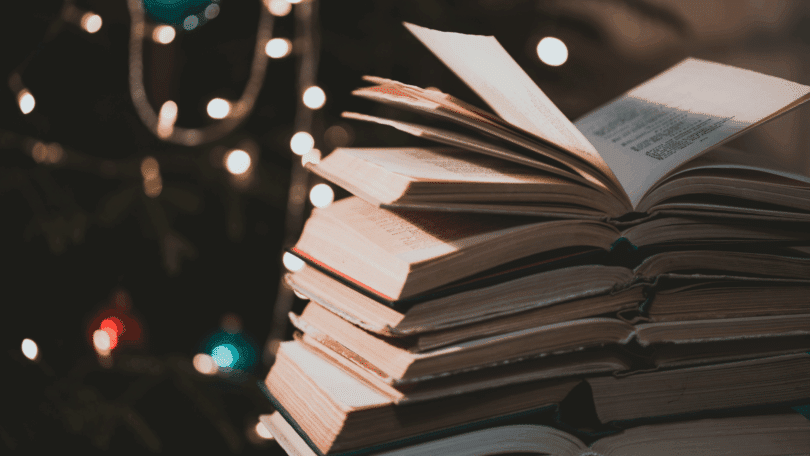 Na imagem, há uma pilha de livros. Ao fundo, há uma árvore de natal.