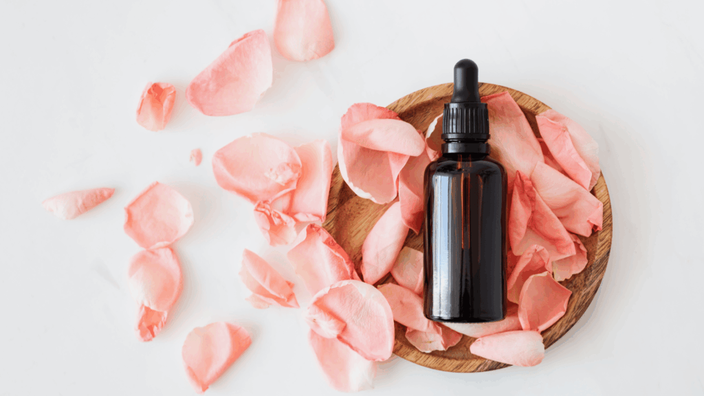 No foco da imagem, há um frasco com óleo essencial. Ao redor, há pétalas de uma flor cor de rosa.
