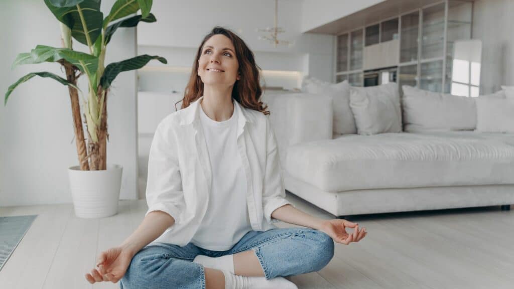 Imagem de uma jovem mulher, sentada no chão de uma sala de ambiente clean, fazendo uma meditação, simbolizando o conceito de paz de espírito e saúde mental.
