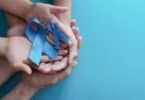 Imagem das mãos de um adulto e de uma criança segurando dois laços de fita na cor azul, simbolizando o Setembro Azul, que é um mês de conscientização e visibilidade para a comunidade surda, dedicado à celebração das suas conquistas e à divulgação da cultura surda e da Libras (Língua Brasileira de Sinais).