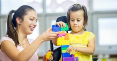 Imagem de uma mãe atípica, brincando com sua filha que precisa de cuidados especiais. Elas estão montando uma peça com jogo de Lego.