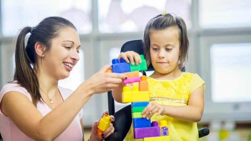 Imagem de uma mãe atípica, brincando com sua filha que precisa de cuidados especiais. Elas estão montando uma peça com jogo de Lego.