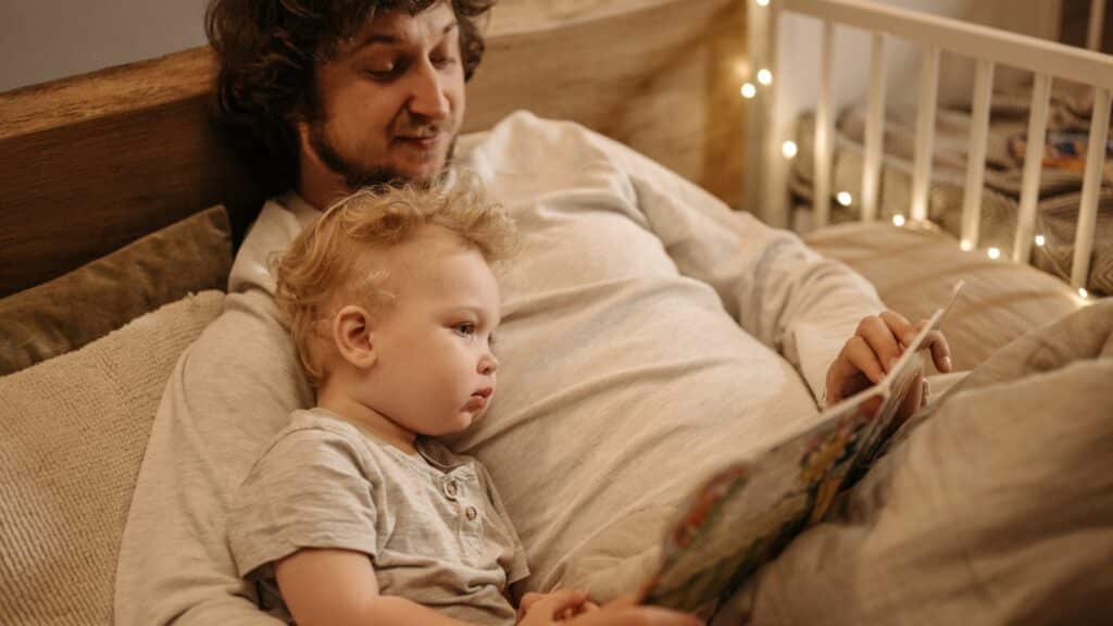 Imagem de um pai lendo um livro de história para o seu filho deitado em sua cama.
