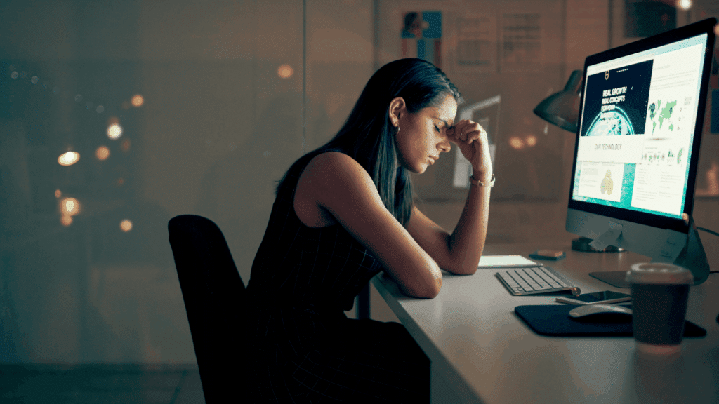 Uma mulher está trabalhando na sua mesa do escritório. Ela está cabisbaixa, com os olhos fechados e apoia uma mão na testa, indicando estar cansada.