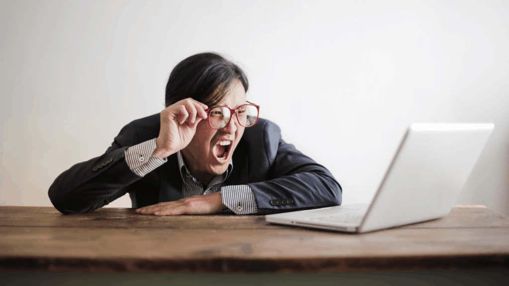 Um homem usando óculos e terno, gritando com expressão de frustração enquanto olha para um notebook sobre a mesa.