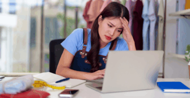 Uma jovem mulher empreendedora está mexendo no seu notebook. Ela apoia uma de suas mãos na cabeça e possui uma expressão de preocupação ou dúvida no rosto, indicando estar preocupada com seu negócio.