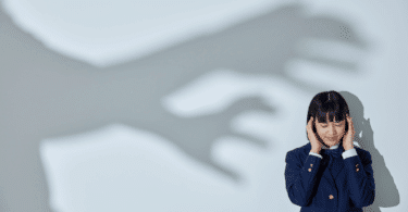 Mulher de olhos fechados e expressão tensa segurando a cabeça, enquanto uma grande sombra de mãos estendidas aparece na parede atrás dela.