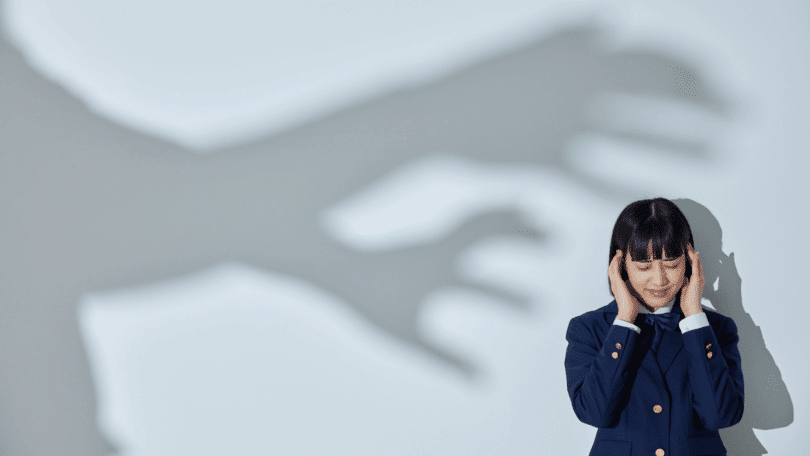 Mulher de olhos fechados e expressão tensa segurando a cabeça, enquanto uma grande sombra de mãos estendidas aparece na parede atrás dela.