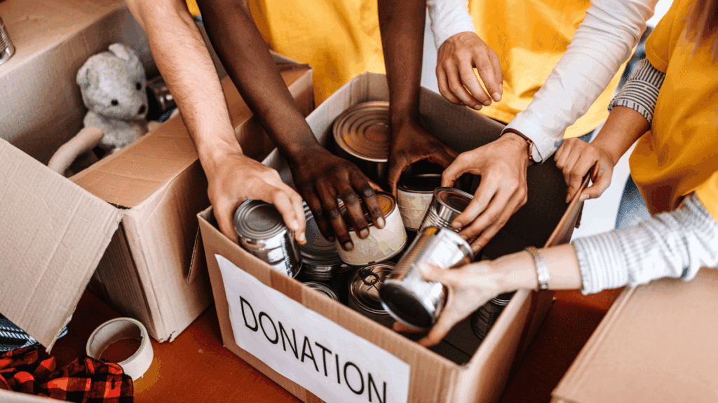 As mãos de várias pessoas colocando alimentos enlatados em uma caixa de doação estão em destaque na imagem.