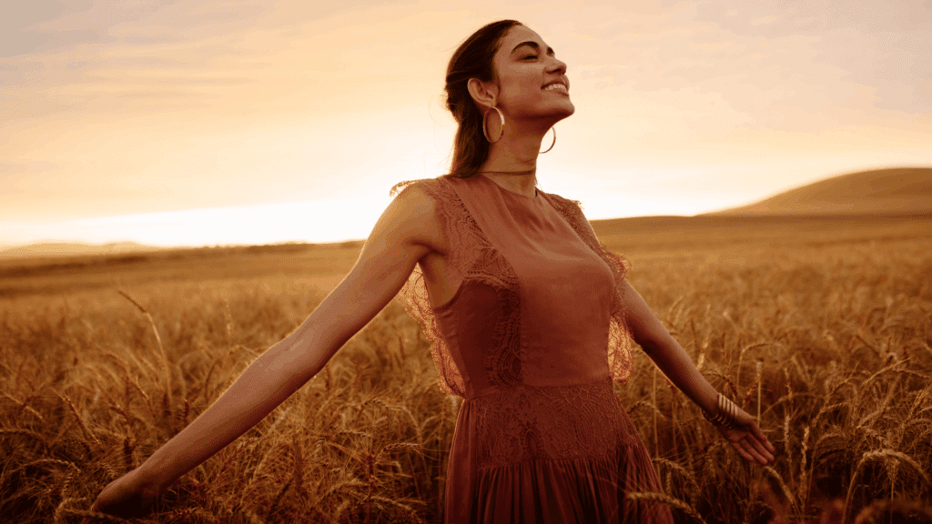 Uma mulher está com os braços abertos, a cabeça levantada e sorrindo. Ela está sentindo a brisa do campo. Ao redor, há plantas, o pôr do sol e a natureza.