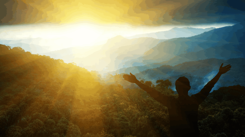 Silhueta de uma pessoa com os braços erguidos em direção ao nascer do sol, diante de uma paisagem montanhosa coberta por vegetação e banhada por luz dourada.