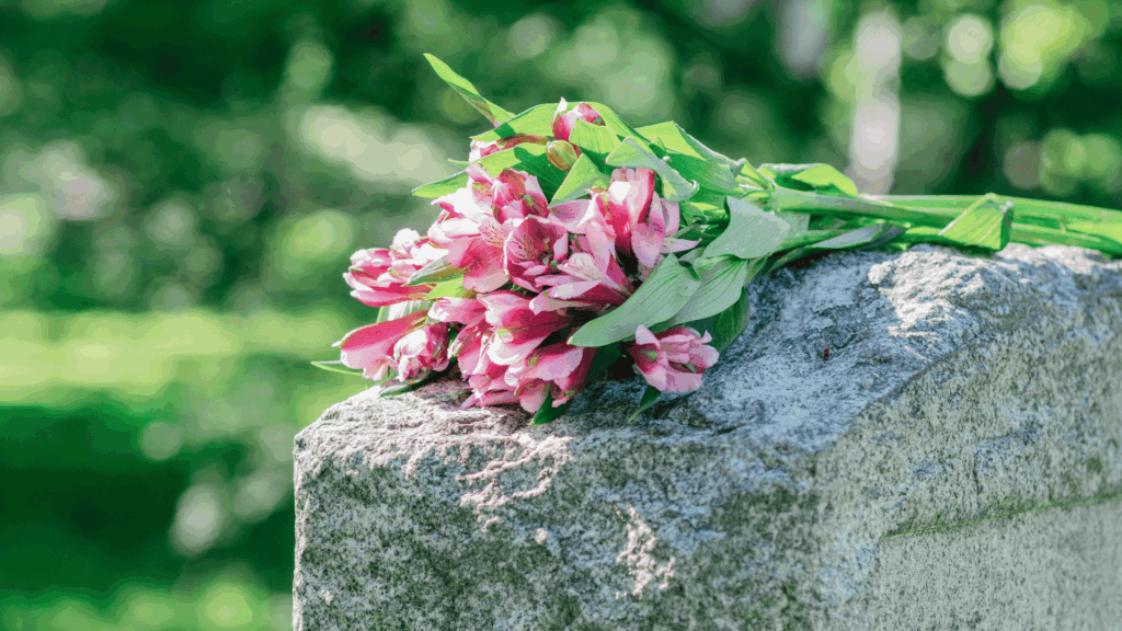 Buquê de flores cor-de-rosa apoiado sobre uma lápide de pedra em um cemitério, com fundo verde desfocado de árvores iluminadas pelo sol.