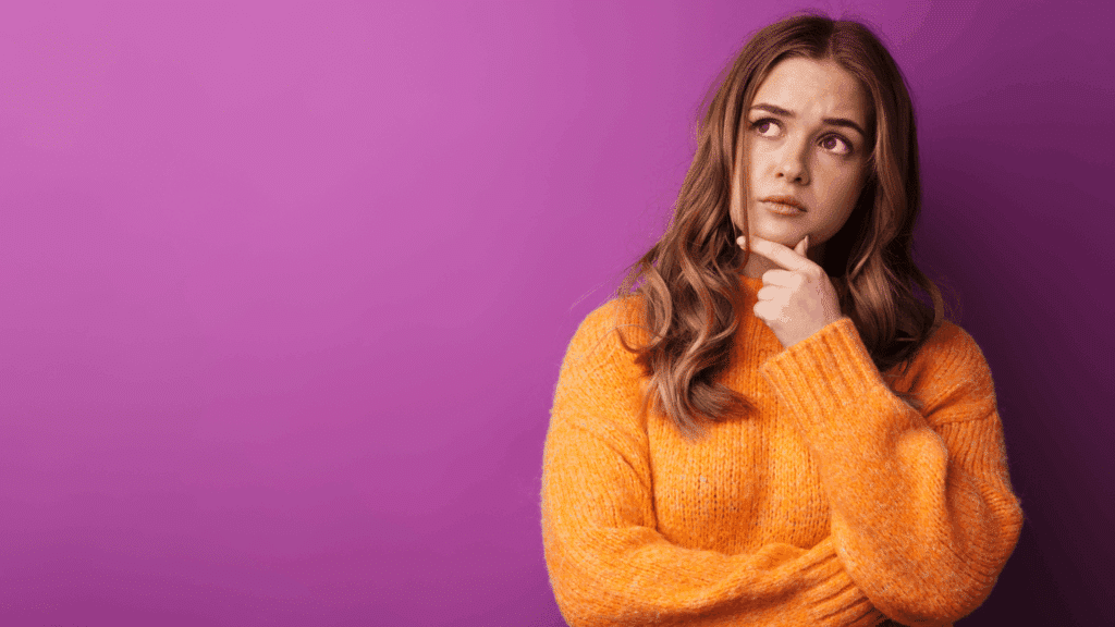 Mulher com expressão pensativa, usando suéter laranja, olhando para cima e tocando o queixo, em frente a um fundo roxo.