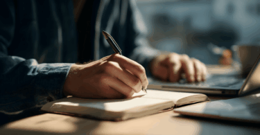 Pessoa escrevendo em um caderno com uma caneta, sentada à mesa com um laptop ao lado, em ambiente iluminado pela luz natural.