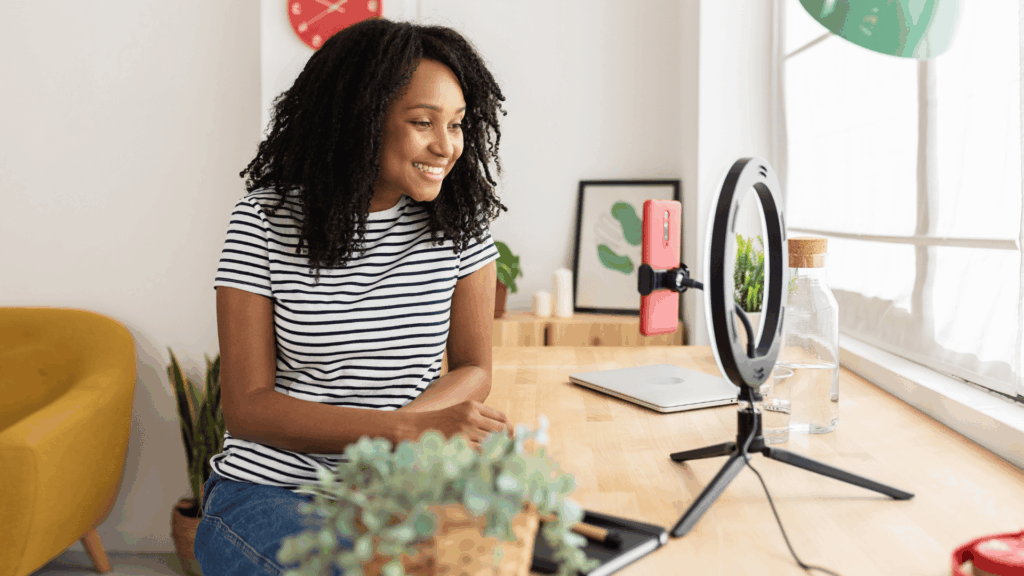 Mulher sorrindo enquanto grava um vídeo com o celular apoiado em um tripé com ring light, sentada à mesa em um ambiente iluminado e aconchegante.
