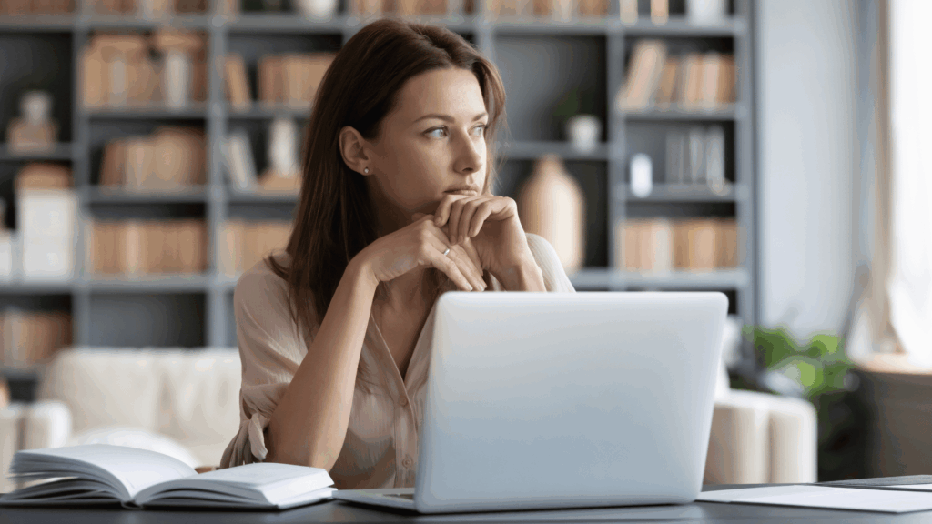 Mulher sentada à mesa com um notebook aberto, olhando pensativa para o lado em um ambiente com estante de livros ao fundo.