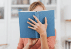 Mulher segurando um livro azul aberto, cobrindo parcialmente o rosto, em um ambiente interno bem iluminado.