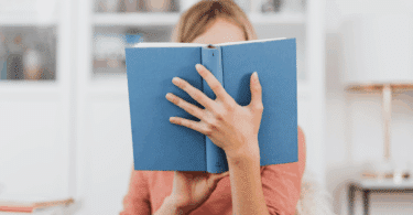 Mulher segurando um livro azul aberto, cobrindo parcialmente o rosto, em um ambiente interno bem iluminado.