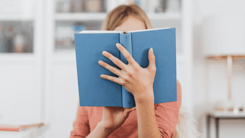 Mulher segurando um livro azul aberto, cobrindo parcialmente o rosto, em um ambiente interno bem iluminado.