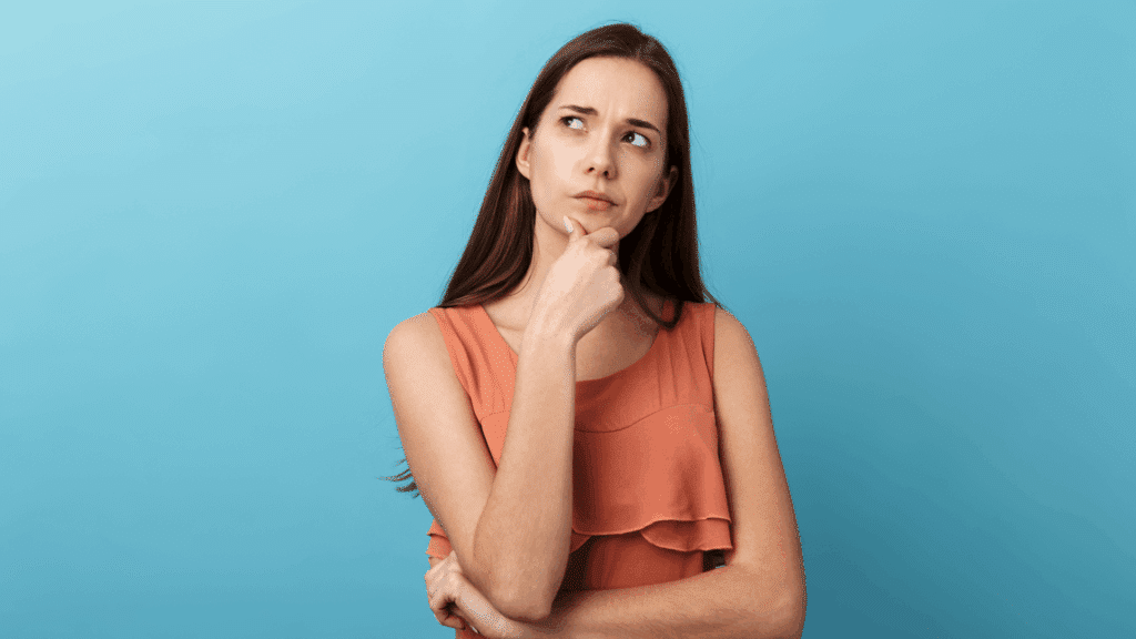 Mulher jovem pensativa diante de um fundo azul, com a mão no queixo e expressão de dúvida.