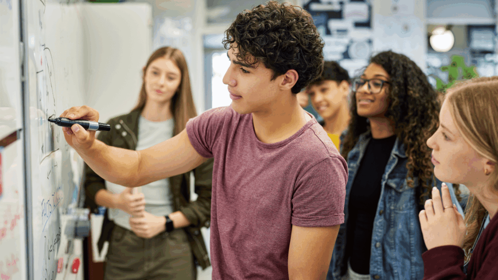 Grupo de estudantes em sala de aula observando um colega escrever em um quadro branco; o aluno à frente segura um marcador e explica algo enquanto os demais assistem com atenção.