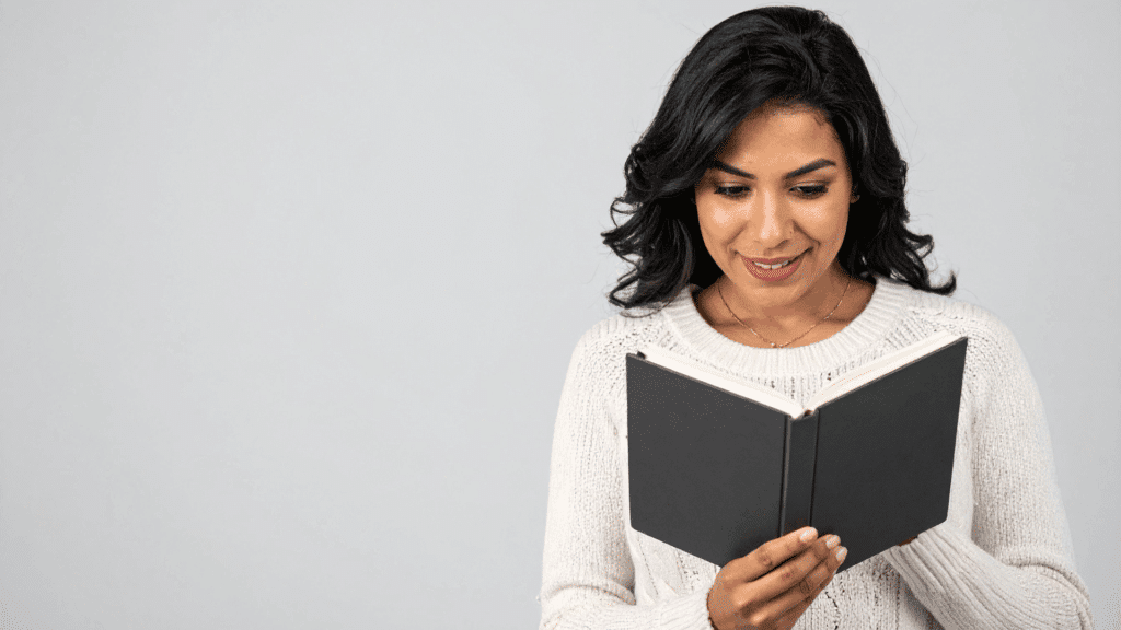 Mulher sorrindo enquanto lê um livro de capa cinza, vestindo um suéter claro e posicionada diante de um fundo cinza neutro.